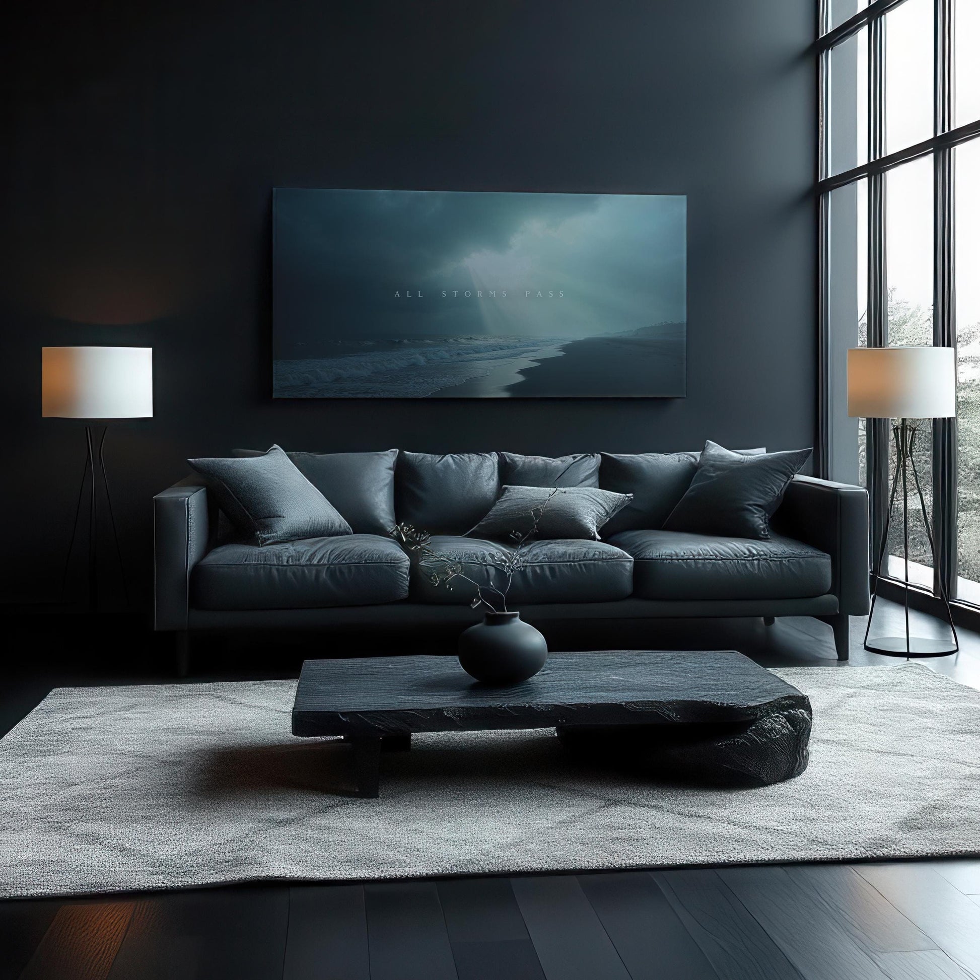 A moody, modern living room with dark walls and a large canvas reading &quot;All Storms Pass&quot; above a black leather sofa. The space includes soft gray cushions, black coffee table, light gray rug, and two tall lamps, with natural light streaming in.
