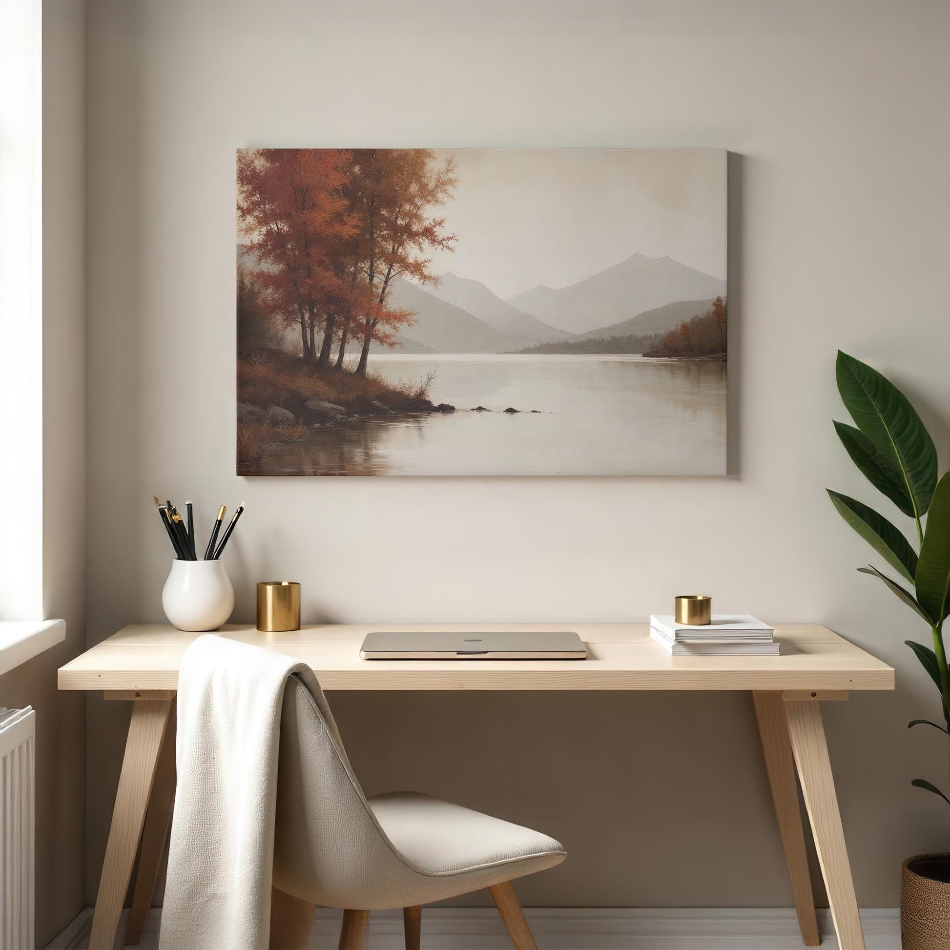 Minimalist workspace featuring a light wood desk, beige chair, and gold-accented decor, including a laptop and potted plant. Above hangs a gallery-wrapped canvas of a serene lake scene with autumn trees and mountains, creating a calming atmosphere.