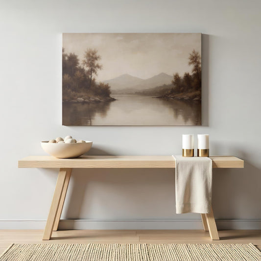 Modern living space with a light wood console table, decorated with a neutral-toned bowl of stones, gold-accented candles, and a beige runner. Above hangs a gallery-wrapped canvas titled Silent Reflection, featuring a serene lake and mountain scene.