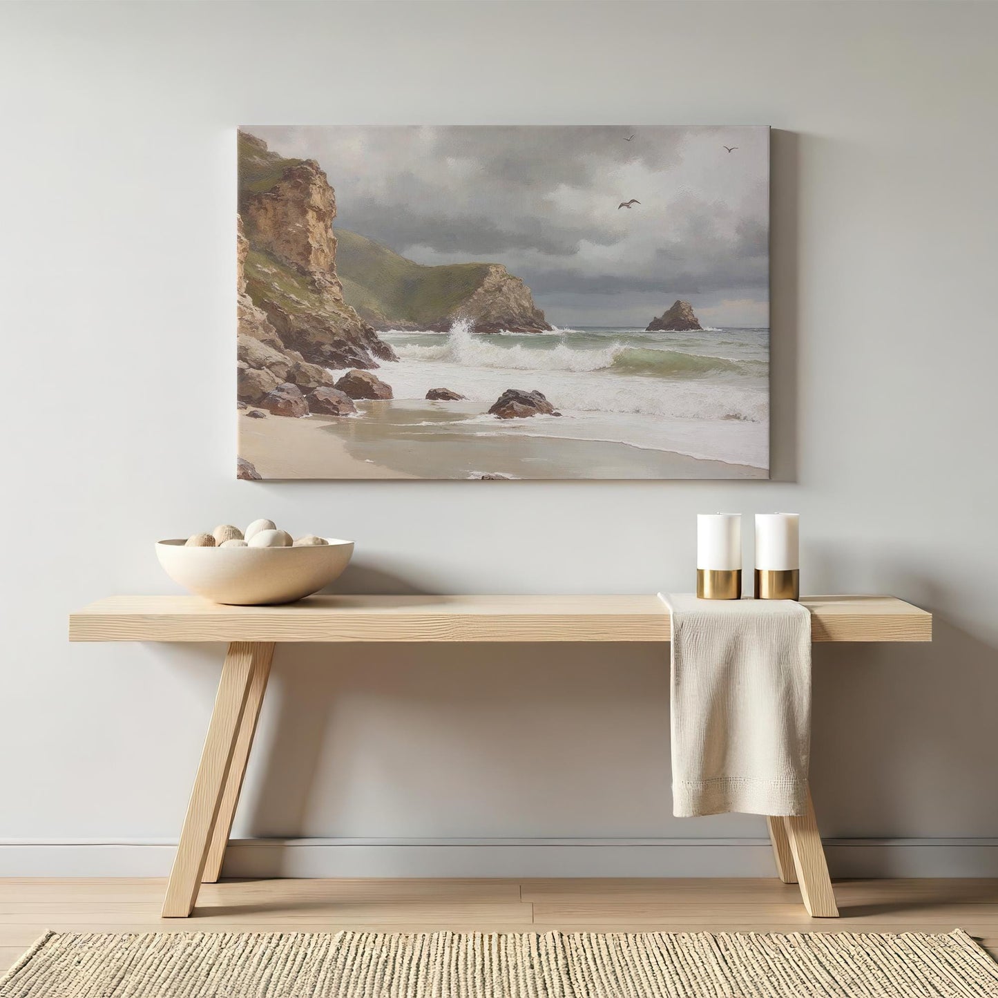 A coastal landscape canvas print featuring a serene beach scene with rocky cliffs, crashing ocean waves, and a cloudy sky. The artwork is displayed above a wooden console table in a minimalist modern room.