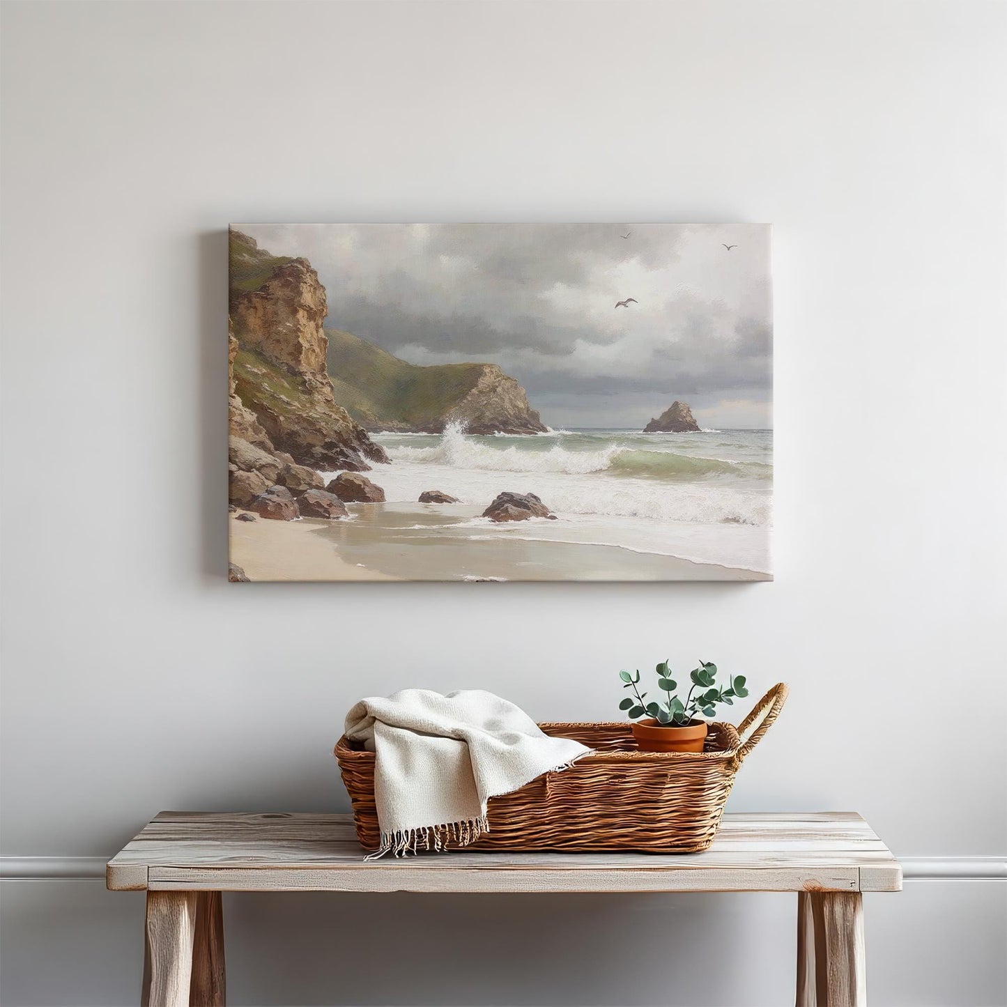 A framed ocean painting featuring rocky cliffs, white-capped waves, and a cloudy coastal sky. The artwork is mounted above a rustic wooden table with a woven basket and cozy decor for a relaxed, beach-inspired aesthetic.