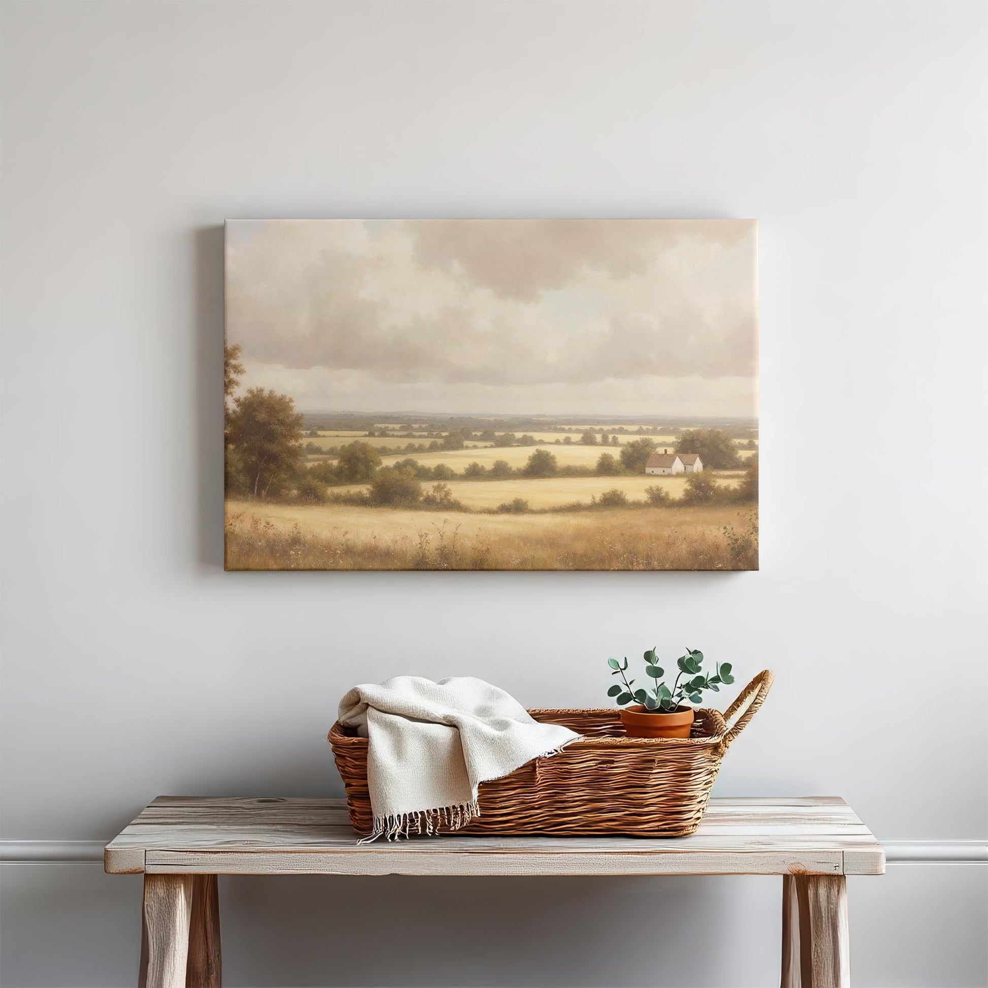 A countryside landscape canvas print displayed above a rustic wooden entryway table, decorated with a woven basket, a light-colored blanket, and a small potted plant. The artwork’s warm tones and pastoral scene create a cozy, welcoming atmosphere.