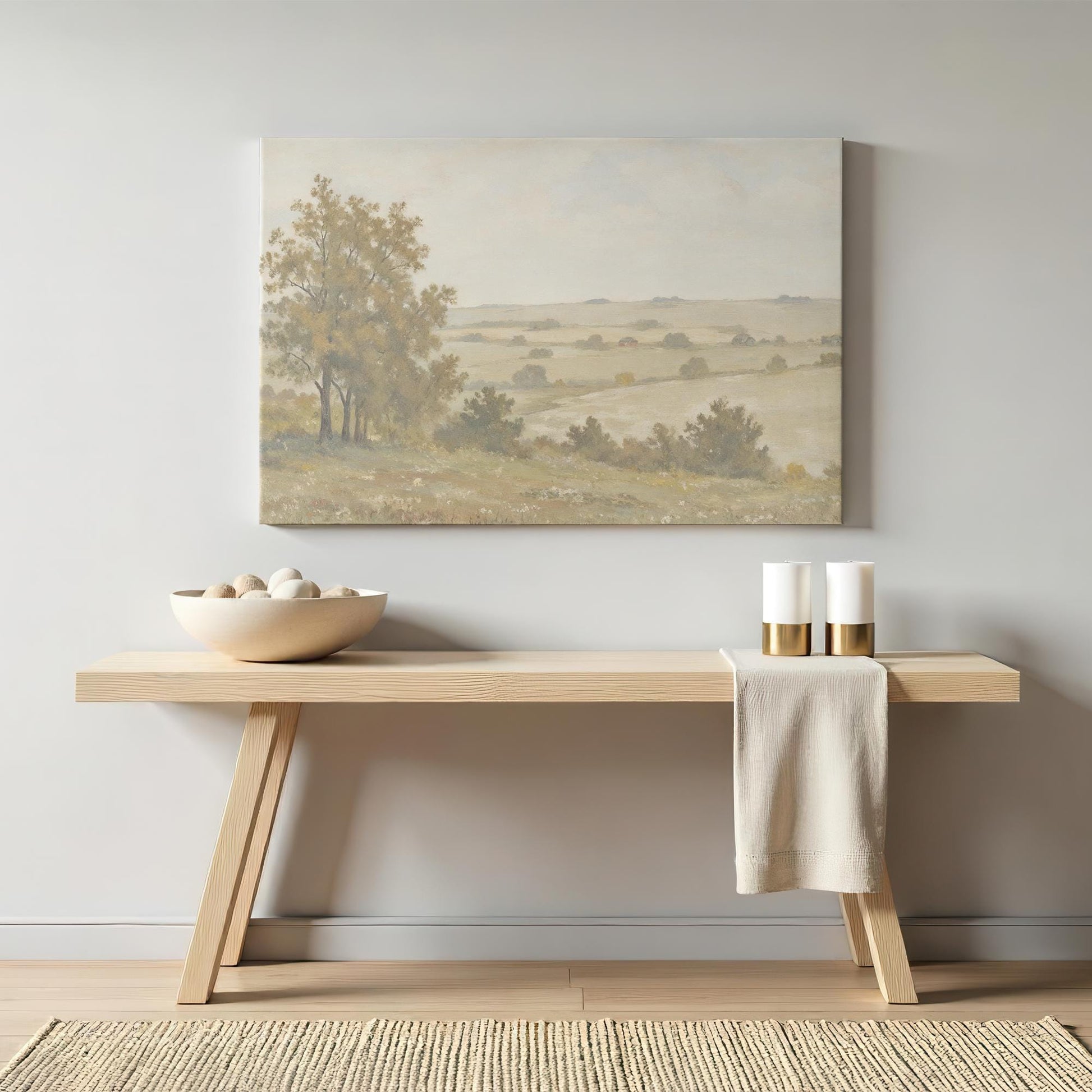 A warm-toned countryside canvas print hanging above a minimalist wooden console table. The soft hues of the artwork complement the neutral decor, with decorative candles and a ceramic bowl enhancing the cozy atmosphere.