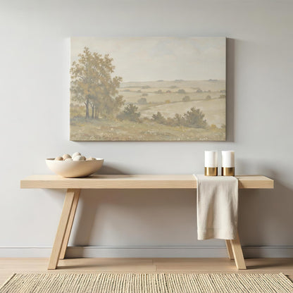 A warm-toned countryside canvas print hanging above a minimalist wooden console table. The soft hues of the artwork complement the neutral decor, with decorative candles and a ceramic bowl enhancing the cozy atmosphere.
