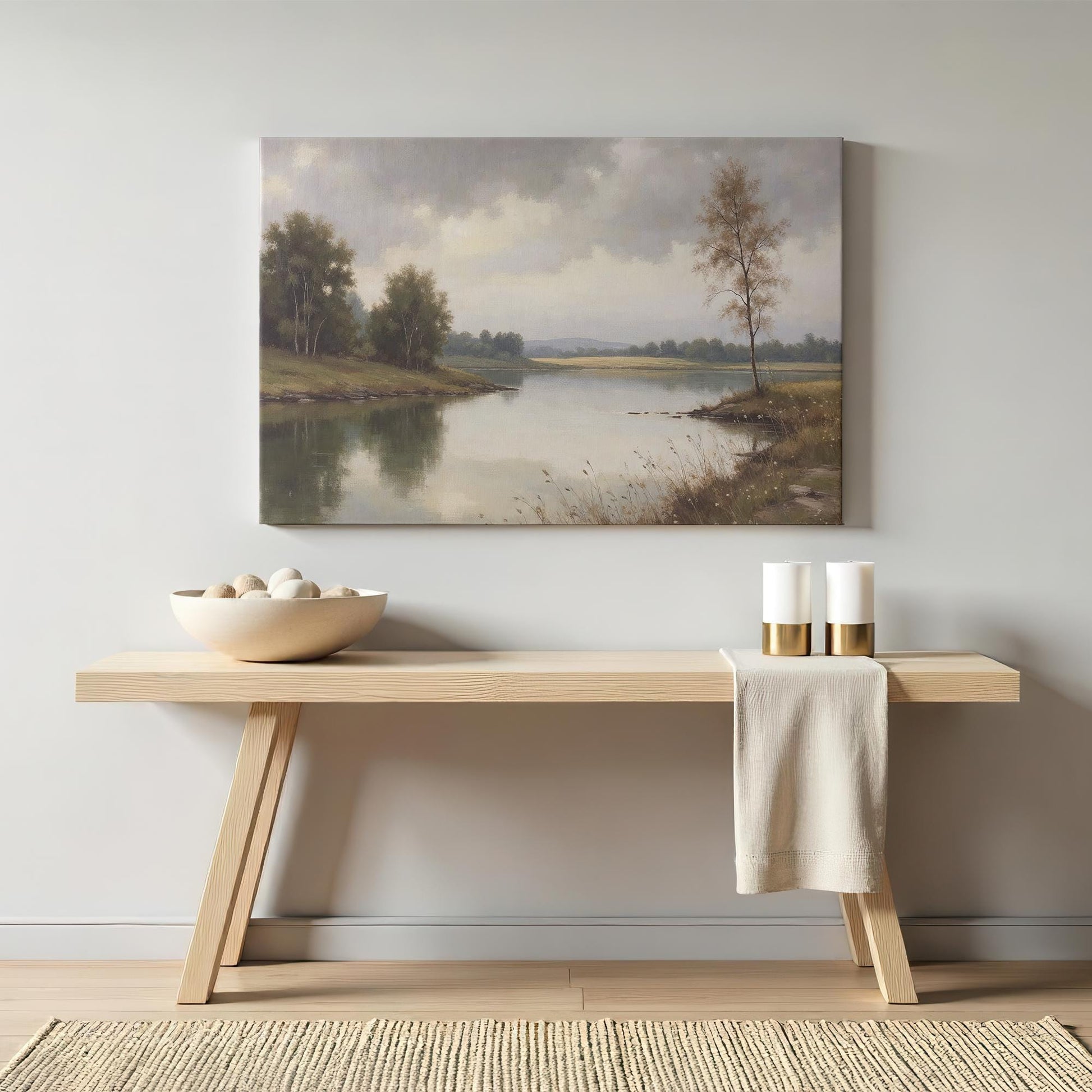 A cozy home interior setting featuring the framed lake landscape canvas above a minimalist wooden console table. The decor includes neutral-colored accents like a woven basket, candles, and soft textiles.