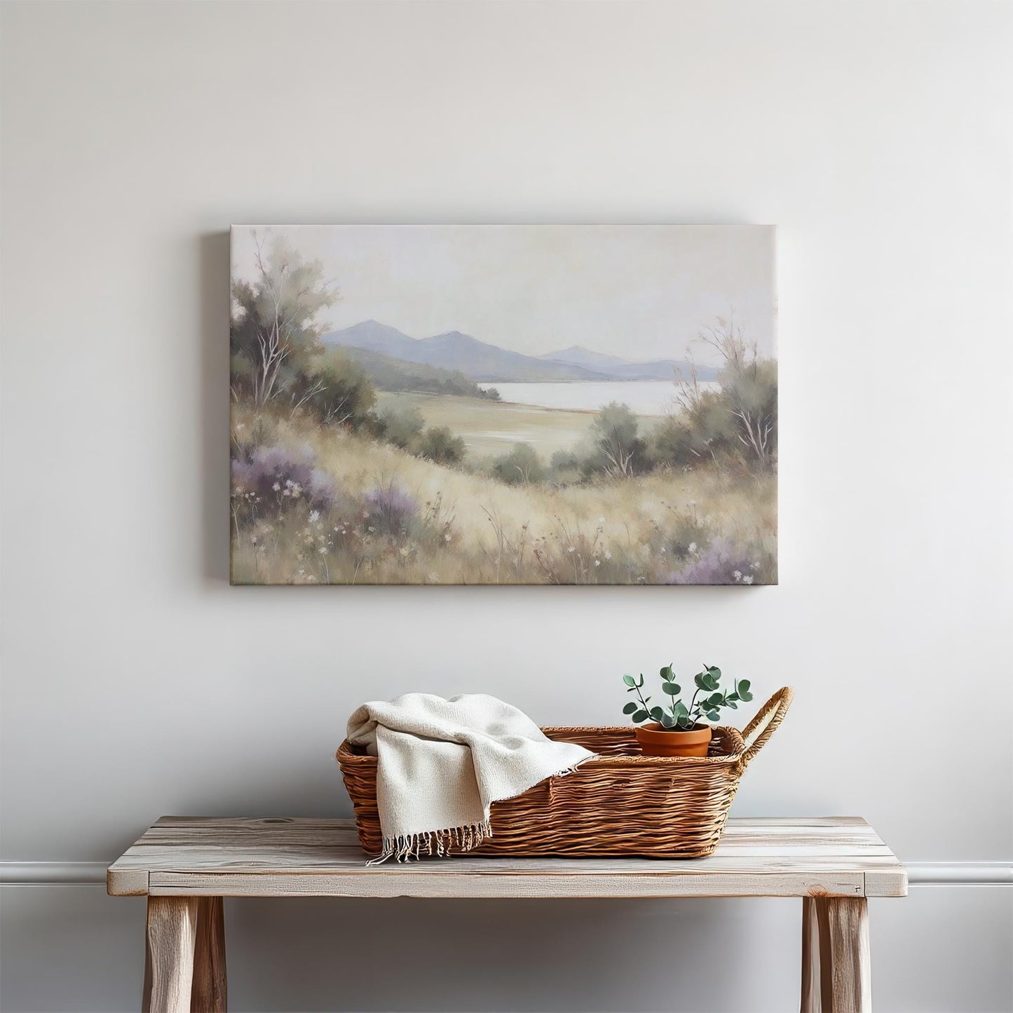 A bright living space with a wood console table styled with neutral decor—ceramic bowl, linen towel, and candles. Above, a landscape canvas of a serene meadow, calm lake, and mountains enhances the room’s natural, minimalist feel.