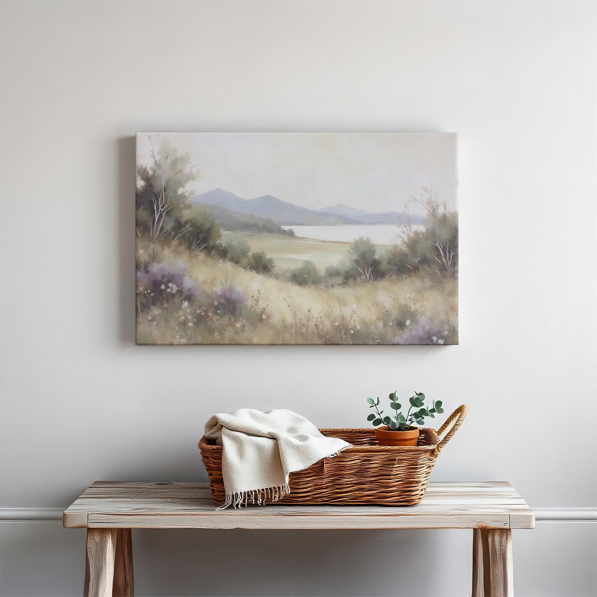 A bright living space with a wood console table styled with neutral decor—ceramic bowl, linen towel, and candles. Above, a landscape canvas of a serene meadow, calm lake, and mountains enhances the room’s natural, minimalist feel.