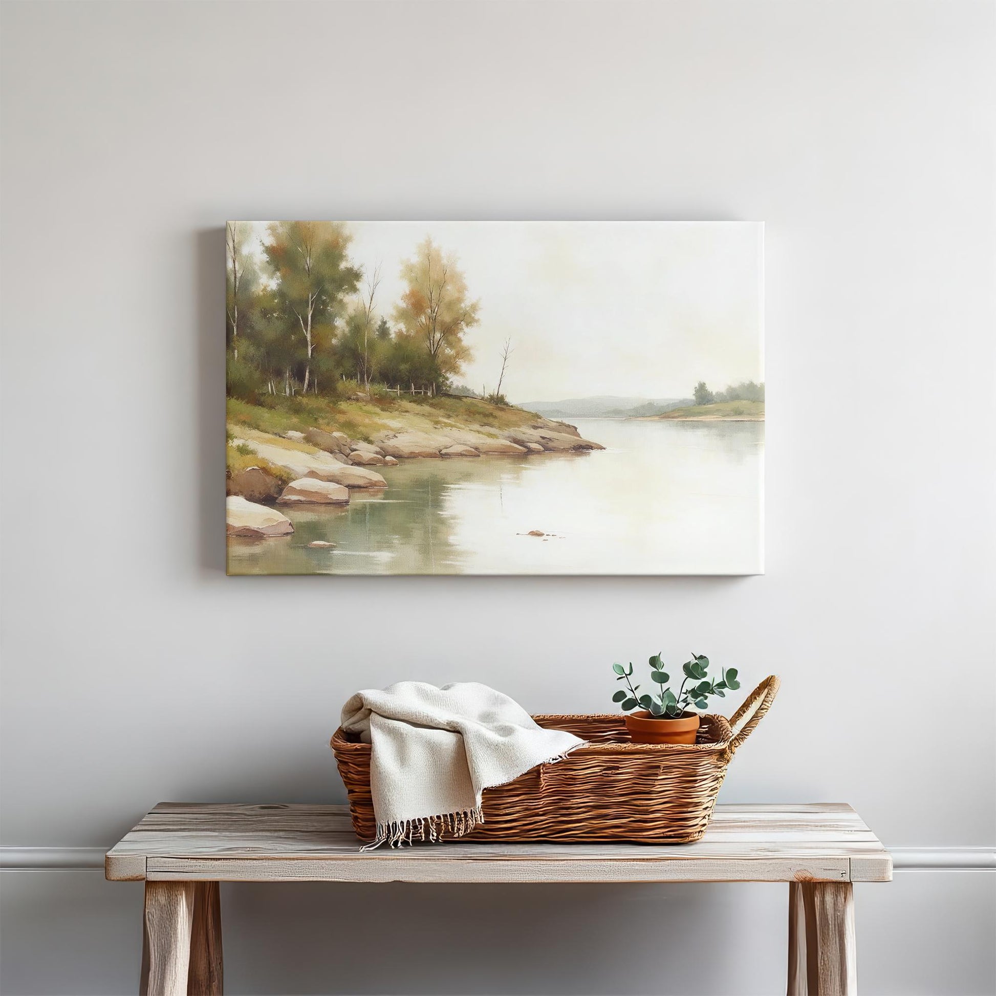 A cozy vignette featuring a framed riverside canvas print mounted on a white wall above a rustic wood console table. A woven basket with a cream throw blanket and a small potted plant add warmth and texture to the minimalist decor.
