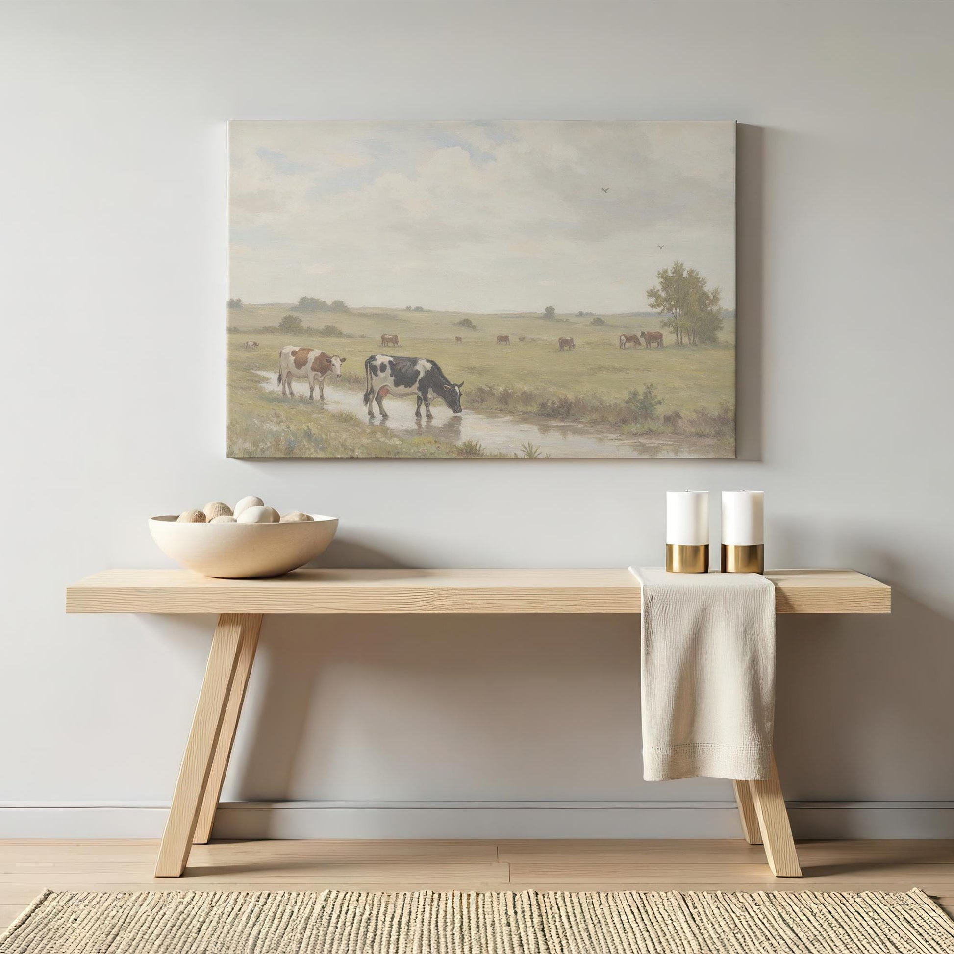 A soft-toned pastoral cow painting displayed above a light wood bench with minimal decor, featuring neutral candles and a ceramic bowl with stones. A cozy, rustic home accent.
