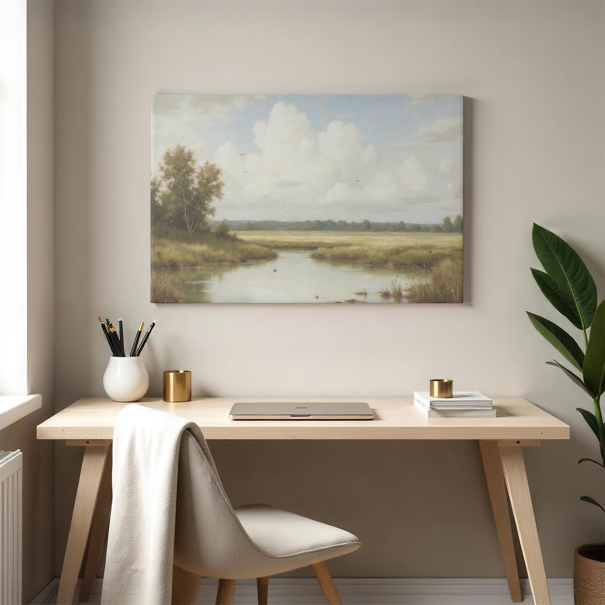 A peaceful countryside landscape canvas featuring a reflective river, lush greenery, and a soft blue sky, mounted above a minimalist wooden desk. The scene is styled with a modern chair, gold-accented decor, and a potted plant.
