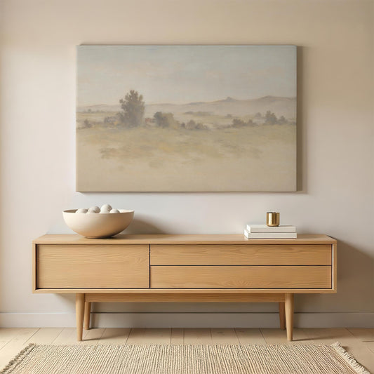 A neutral-toned landscape canvas painting hangs above a minimalist wooden sideboard. A ceramic bowl with decorative stones and a small stack of books complement the modern, serene decor in a well-lit space.