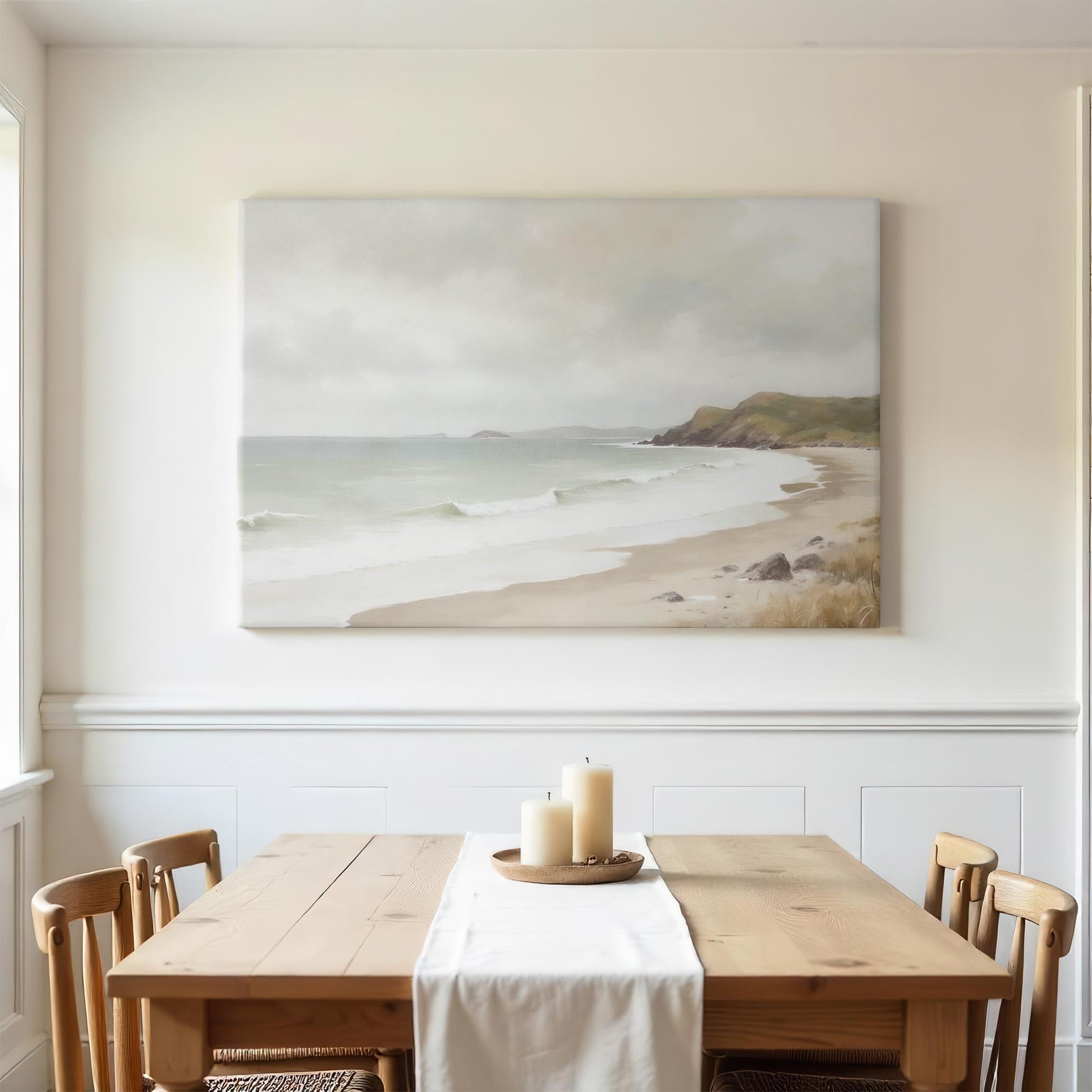 Large coastal canvas art above wooden dining table with white runner. Neutral decor, candle centerpiece, and wooden chairs create a serene and inviting farmhouse dining space with ocean-inspired decor.