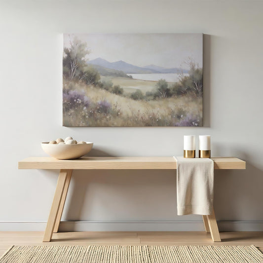 A modern entryway with a wooden console table, woven basket, soft throw, and potted plant. A calming landscape canvas of rolling hills, wildflowers, and a lake with distant mountains adds a serene touch to the neutral decor.