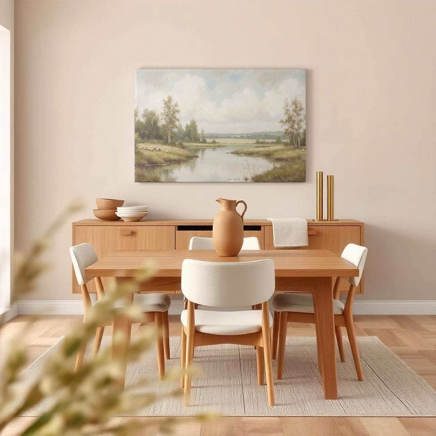 A warm, modern dining room features a wooden table, cream-colored chairs, and a neutral-toned sideboard. Above, a tranquil landscape canvas print of a river and countryside brings nature into the space.