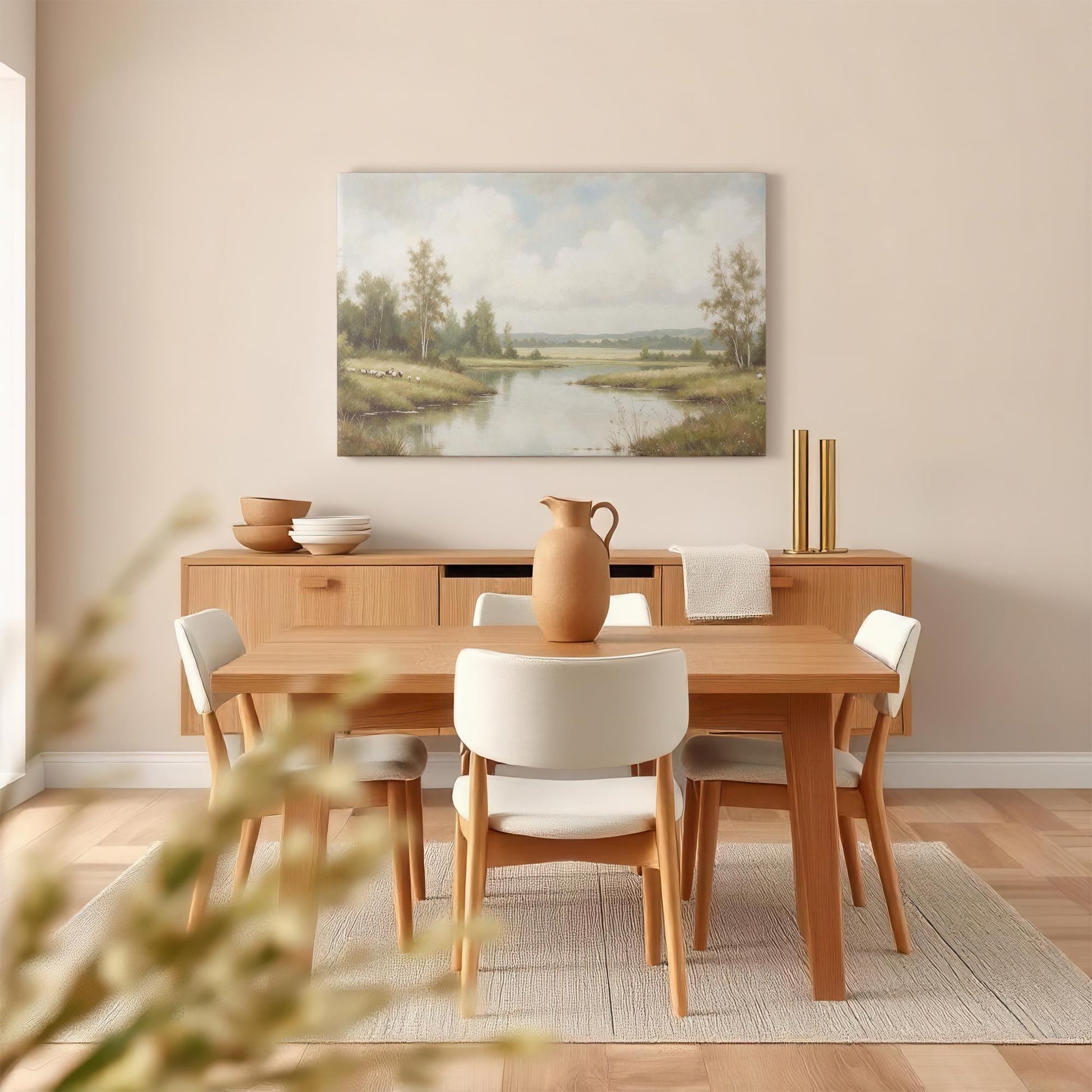 A warm, modern dining room features a wooden table, cream-colored chairs, and a neutral-toned sideboard. Above, a tranquil landscape canvas print of a river and countryside brings nature into the space.