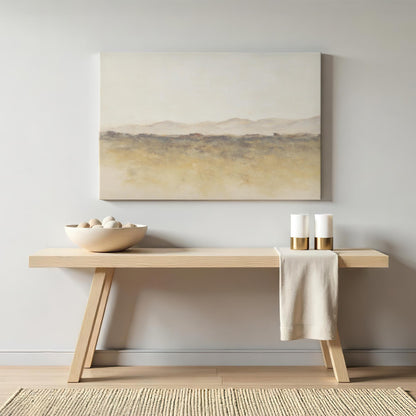 A warm-toned wooden console table against a neutral wall, adorned with a soft abstract landscape canvas. The table holds a bowl of stones and gold-accented candles for a simple, elegant touch.