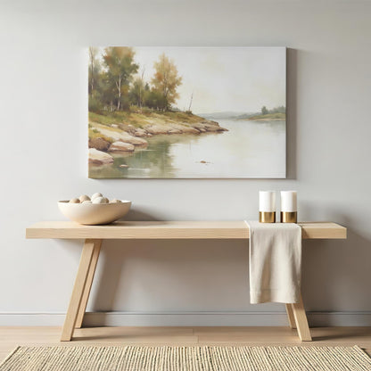 A framed canvas painting of a tranquil riverside landscape with lush trees and smooth rocks, hung on a light gray wall above a wooden bench. The bench has a neutral-toned bowl with decorative stones and two white candles with gold accents.