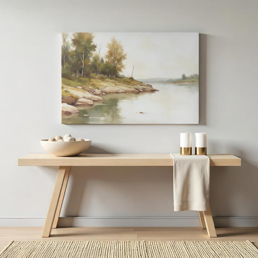 A framed canvas painting of a tranquil riverside landscape with lush trees and smooth rocks, hung on a light gray wall above a wooden bench. The bench has a neutral-toned bowl with decorative stones and two white candles with gold accents.