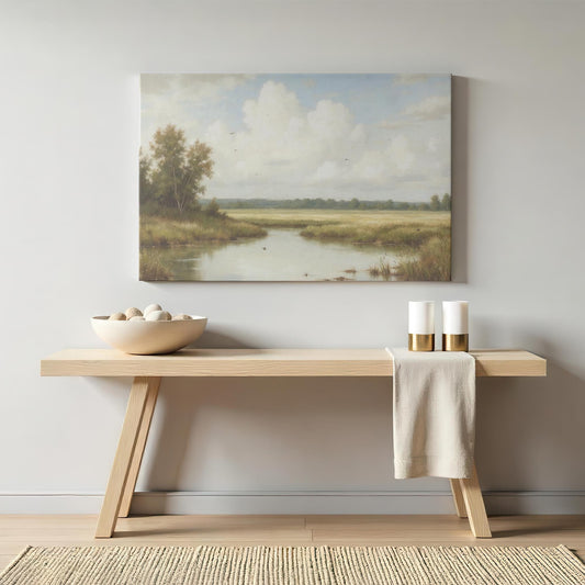A large canvas print of a serene landscape featuring a calm river, grassy fields, and a soft blue sky with fluffy clouds. Displayed above a modern wooden console table with a decorative bowl of stones and two elegant candle holders.