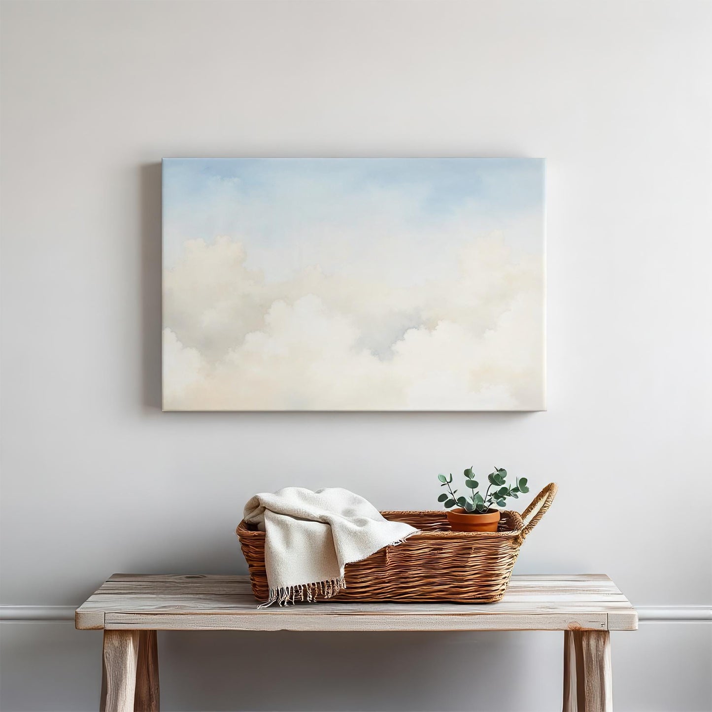 Serene cloudscape canvas print styled in a minimalist home setting. A woven basket with a cozy throw and greenery complements the light, airy aesthetic, creating a peaceful entryway or living space.