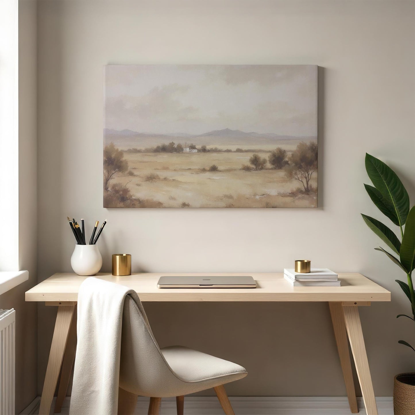 A neutral-hued canvas print of a tranquil desert landscape with rolling hills and a small house, complementing a modern home office space with a wooden desk, gold accents, and soft natural lighting.