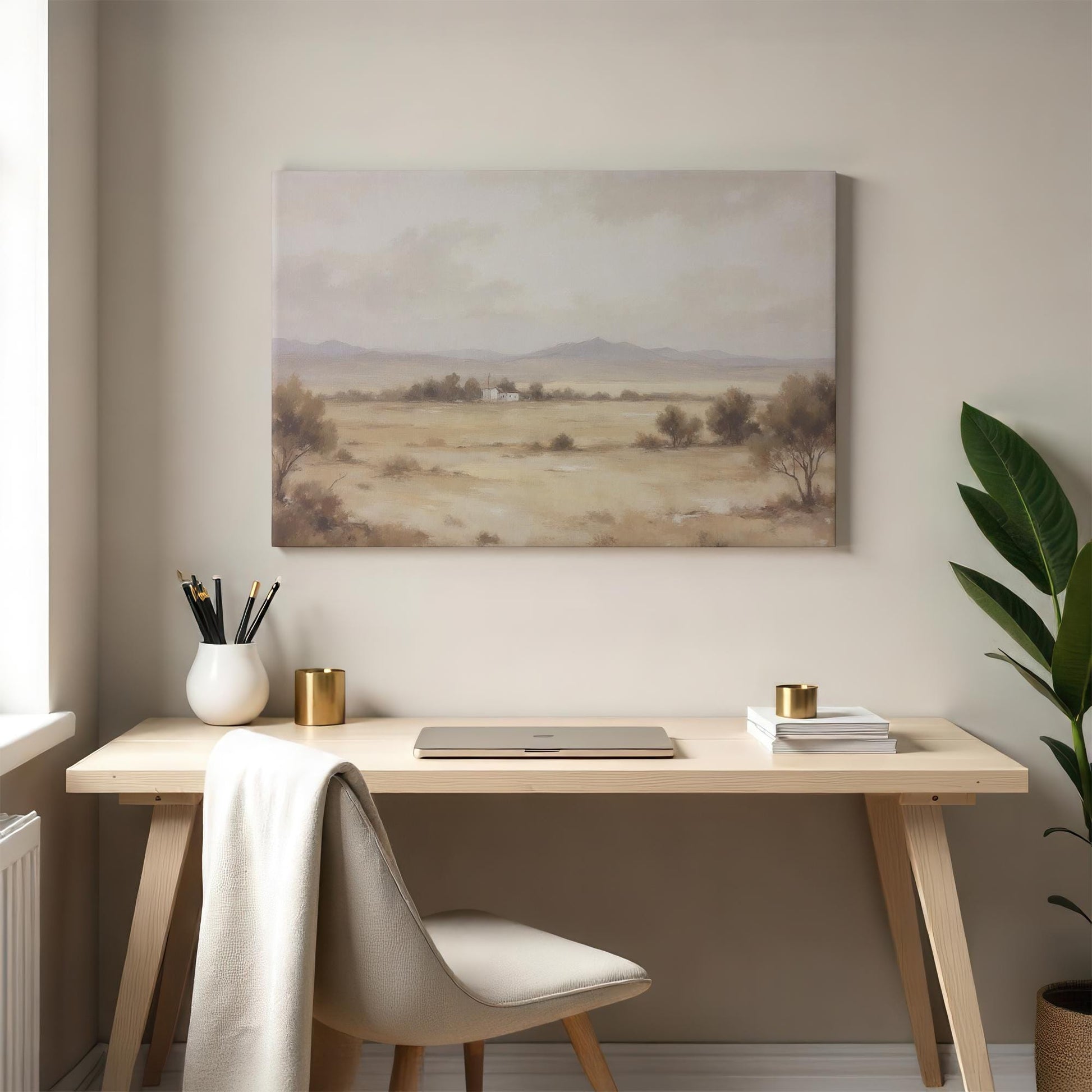 A neutral-hued canvas print of a tranquil desert landscape with rolling hills and a small house, complementing a modern home office space with a wooden desk, gold accents, and soft natural lighting.