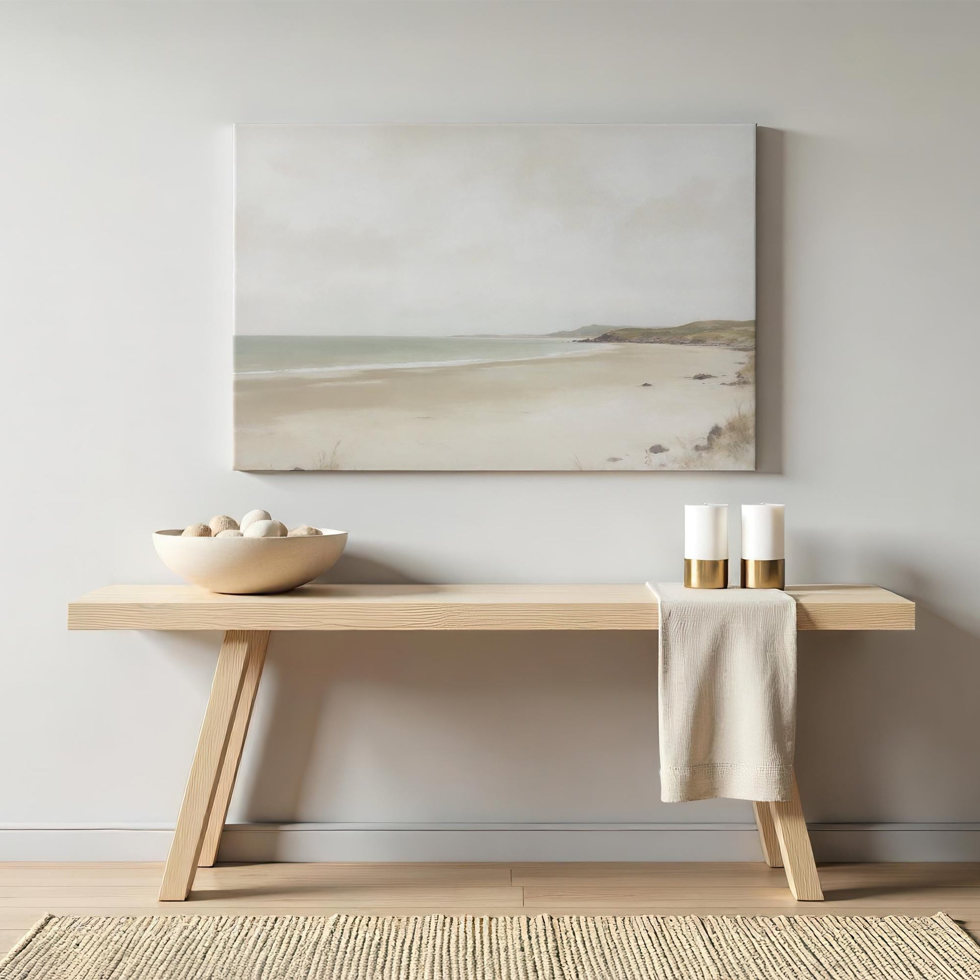 Neutral coastal artwork of a serene beach scene in soft muted tones, styled above a minimalist wooden console table. The decor features a modern, organic aesthetic with ceramic accents and gold candle holders.