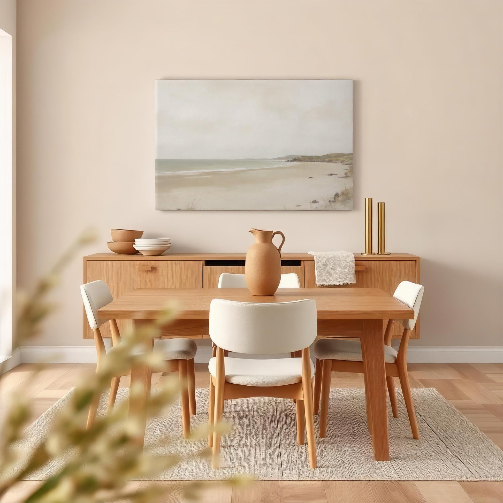 A coastal-inspired dining room with a light wood table and white upholstered chairs. Above the sideboard, a large neutral-toned beach landscape canvas adds a calm, airy feel to the space. A ceramic pitcher centerpiece enhances the aesthetic.