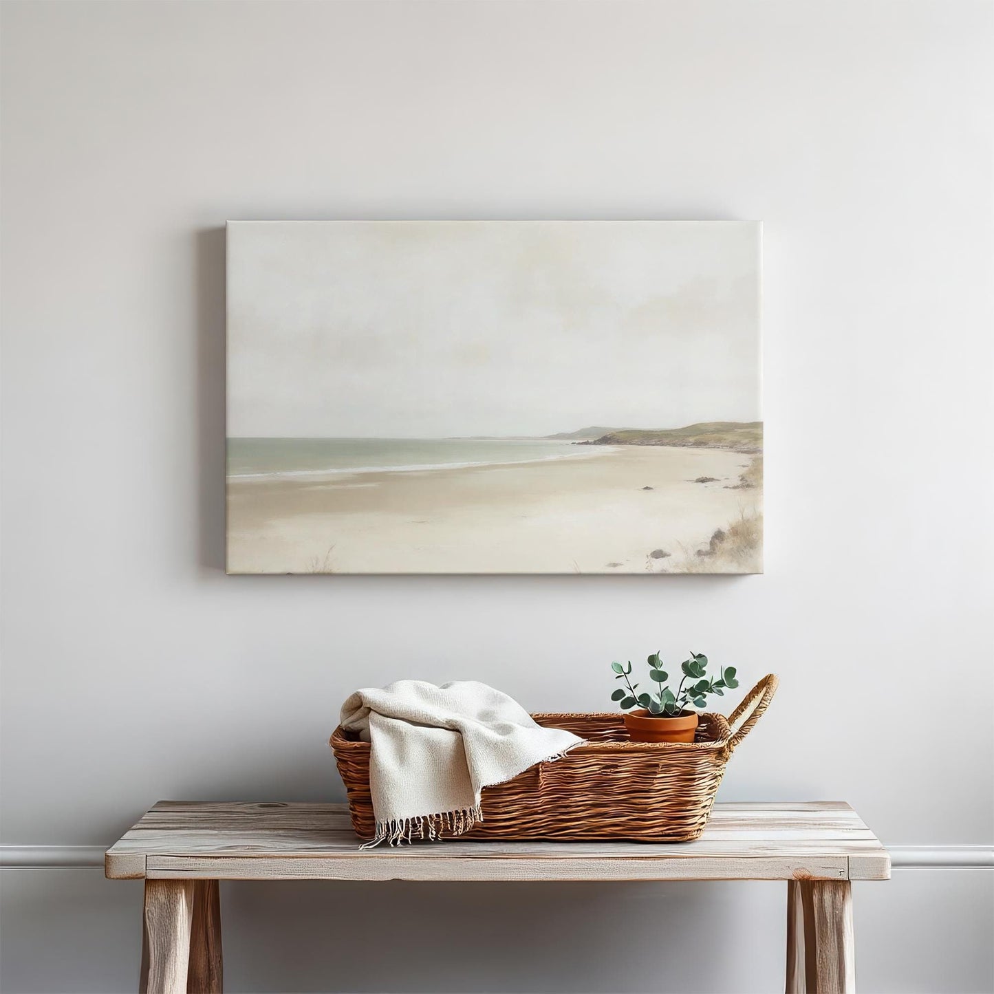 Minimalist coastal wall art featuring a serene seascape canvas with soft neutral tones. Displayed above a wooden console table styled with a woven basket, a cozy throw, and a potted plant in a modern, coastal-inspired home.