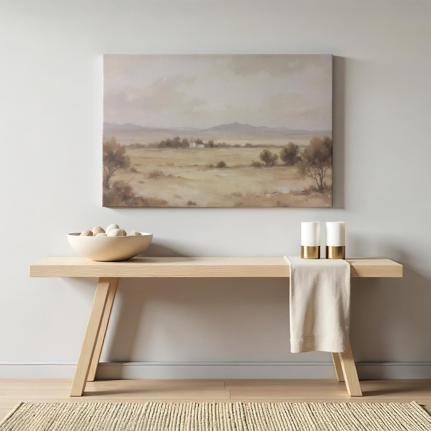 A neutral-toned canvas print of a serene desert landscape with distant mountains and a small farmhouse, styled above a minimalist wooden console table with modern decor, candles, and a ceramic bowl.