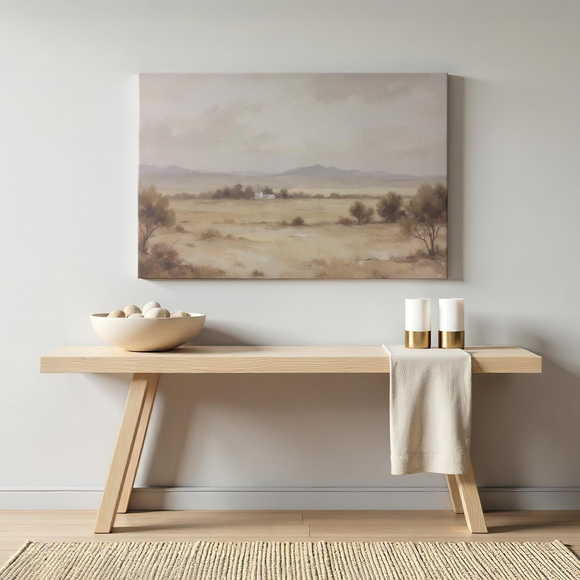A neutral-toned canvas print of a serene desert landscape with distant mountains and a small farmhouse, styled above a minimalist wooden console table with modern decor, candles, and a ceramic bowl.