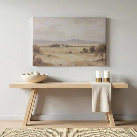 A neutral-toned canvas print of a serene desert landscape with distant mountains and a small farmhouse, styled above a minimalist wooden console table with modern decor, candles, and a ceramic bowl.