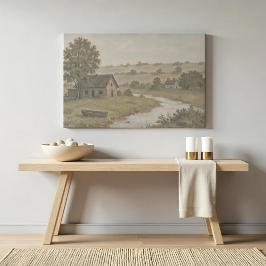 A cozy entryway featuring a light wood console table adorned with a ceramic bowl of stones and candles, with a vintage countryside landscape canvas showcasing a river, rustic home, and rolling green hills hanging above.