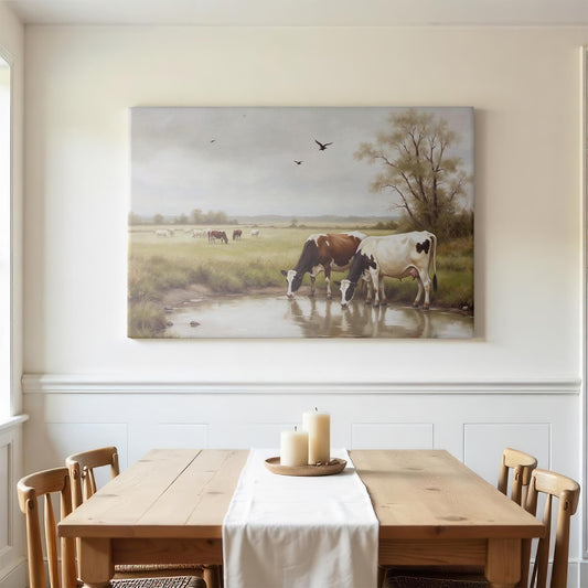 A farmhouse-style dining room featuring a large canvas painting of cows drinking from a pond in a pastoral field. The wooden dining table with candles and natural decor creates a warm, rustic ambiance.