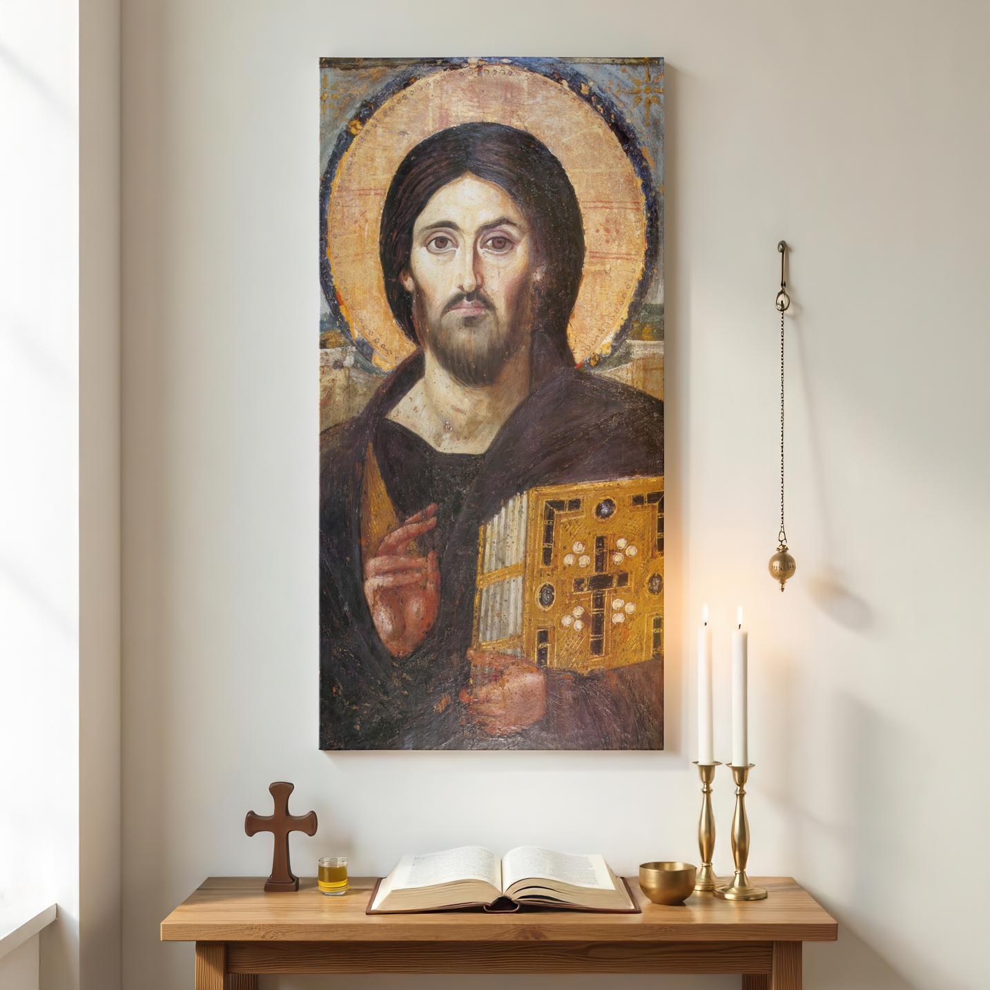 A tall canvas featuring the Icon of Christ Pantocrator hangs in an Orthodox prayer corner. Below, a wooden table holds an open Bible, a wooden cross, a brass bowl, and three white candles in a gold holder, creating a sacred space.