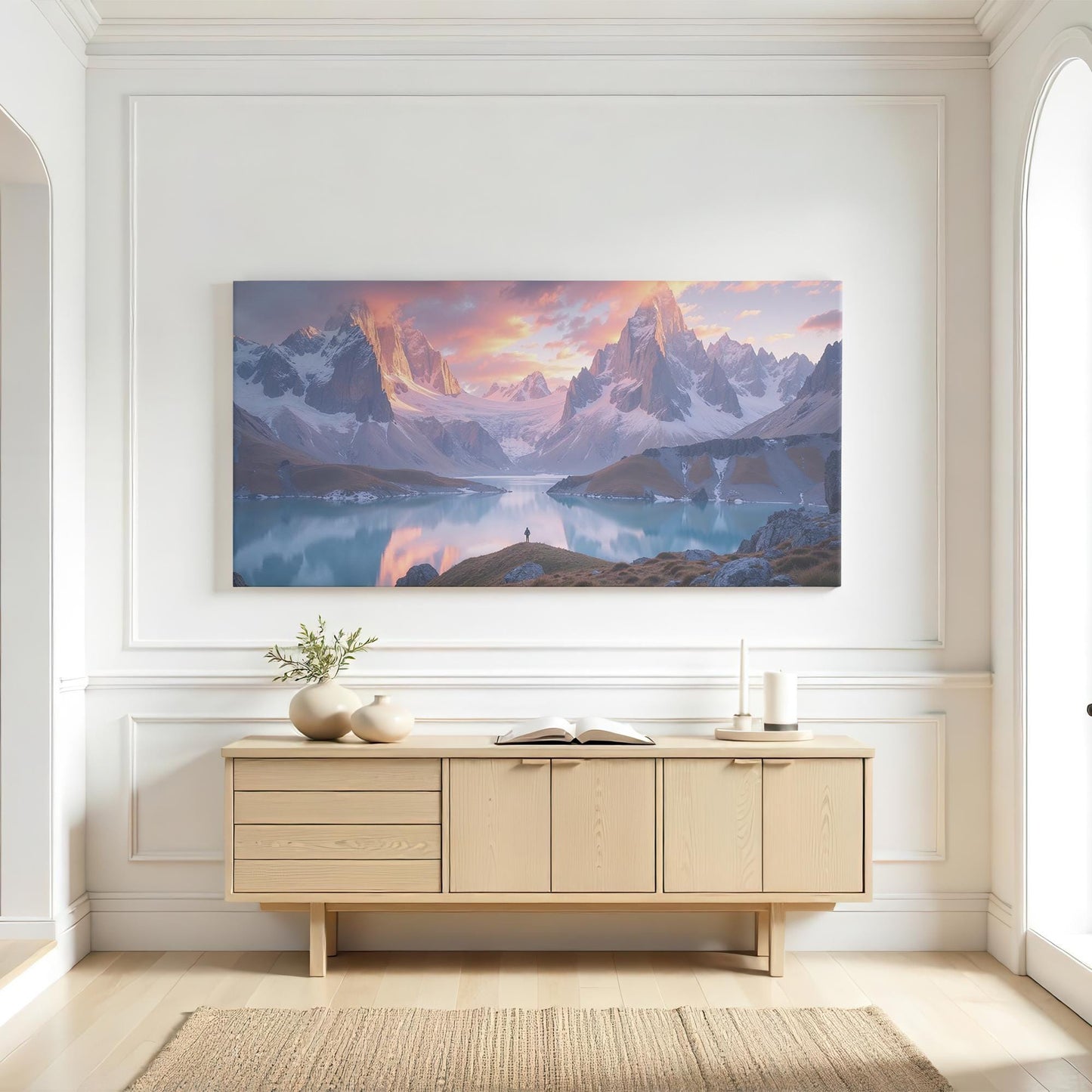 A large panoramic mountain wall art piece displayed in a modern living room with dark interior tones. The artwork showcases snow-capped peaks under a pink and orange sunset sky, their reflection shimmering in the still waters of an alpine lake. The dramatic light and shadow bring a sense of depth and awe, creating a luxurious and inspiring ambiance for any space designed for relaxation and contemplation.