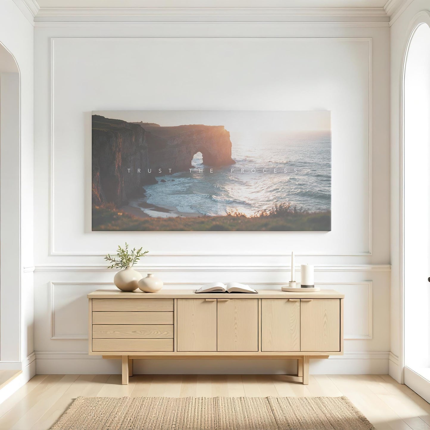 A bright, airy living space with white paneled walls and a modern wooden console table. Above it, a large canvas print of a coastal cliff bathed in sunset light with the words &quot;Trust the Process&quot; adds a motivational touch. The space is styled with neutral ceramic vases, a small plant, and an open book. Natural light pours in, enhancing the warm, serene aesthetic of the room.