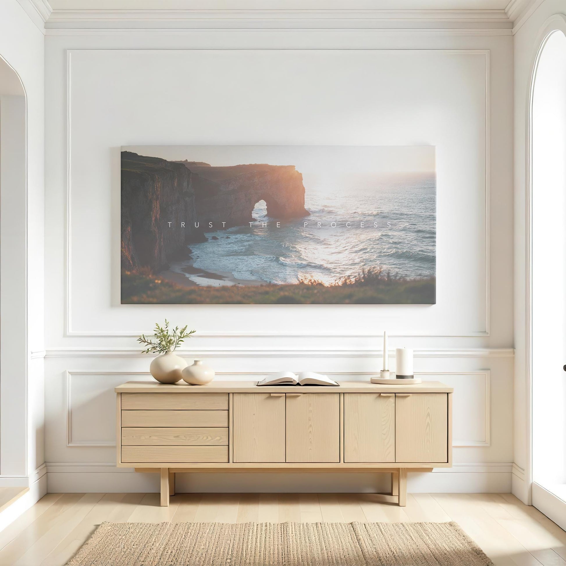 A bright, airy living space with white paneled walls and a modern wooden console table. Above it, a large canvas print of a coastal cliff bathed in sunset light with the words &quot;Trust the Process&quot; adds a motivational touch. The space is styled with neutral ceramic vases, a small plant, and an open book. Natural light pours in, enhancing the warm, serene aesthetic of the room.