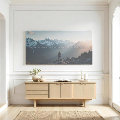 A minimalist white living space with a large inspirational mountain canvas centered above a light wood console table. The artwork captures a lone hiker standing in awe of a breathtaking sunrise-lit mountain range, with the quote &quot;The best view comes from the hardest climb&quot;. Neutral-toned ceramic vases, an open book, and soft natural light enhance the peaceful and modern aesthetic.
