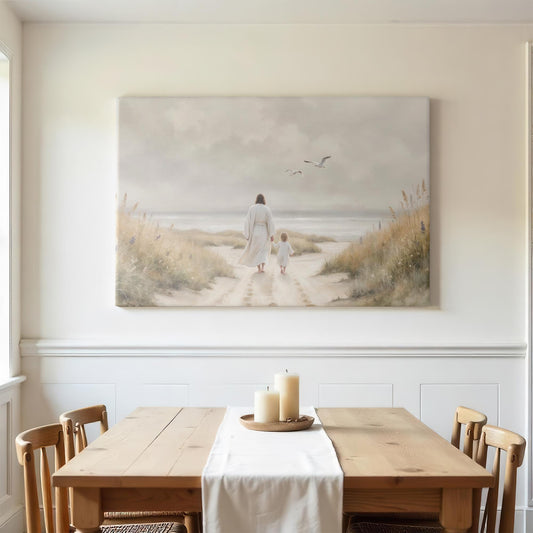 A bright farmhouse-style dining room with a large canvas of Jesus and a child walking toward the ocean. The wooden dining table has a white linen runner and a tray with pillar candles. Natural wood chairs and white walls with wainscoting create a clean, elegant space. Soft daylight enhances the peaceful coastal artwork, making the room feel calm and inviting.