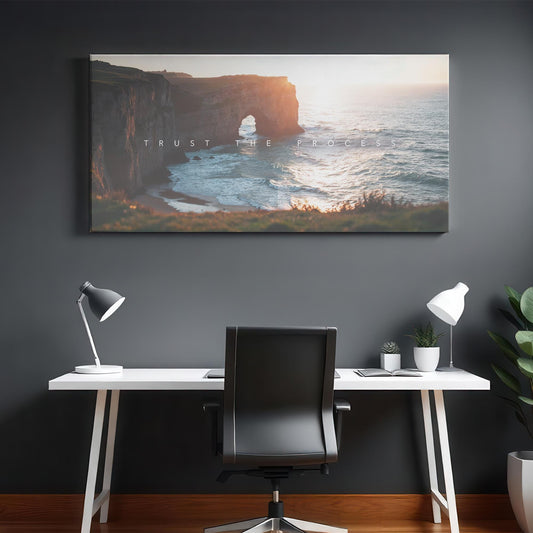 A sleek home office with a dark gray wall features a large canvas print of a coastal cliff at sunset, with &quot;Trust the Process&quot; text. The minimalist white desk has a black leather chair, two modern desk lamps, and small potted plants. A wooden floor and a neutral rug add warmth to the space. The artwork’s golden light and ocean waves create a calming yet motivational atmosphere.