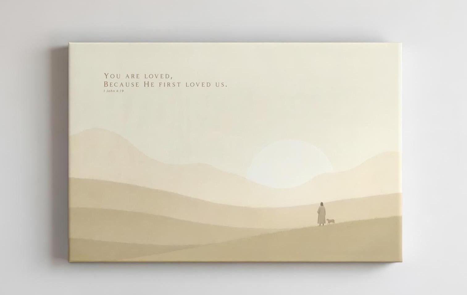 Faith-based wall art with the verse &#39;You are loved, because He first loved us&#39; from 1 John 4:19, displayed above a minimalist light wood desk. The soft, warm-toned canvas features a peaceful pastoral scene with a distant figure and a lamb, creating a calming atmosphere ideal for a nursery, home office, or devotional space.