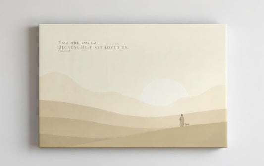 Faith-based wall art with the verse &#39;You are loved, because He first loved us&#39; from 1 John 4:19, displayed above a minimalist light wood desk. The soft, warm-toned canvas features a peaceful pastoral scene with a distant figure and a lamb, creating a calming atmosphere ideal for a nursery, home office, or devotional space.